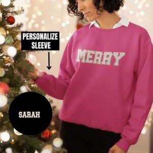May include: A magenta sweatshirt with the word "MERRY" in white block letters. The text "PERSONALIZE SLEEVE" is above a black circle with the name "SARAH" in white. The sweatshirt is a festive holiday garment.