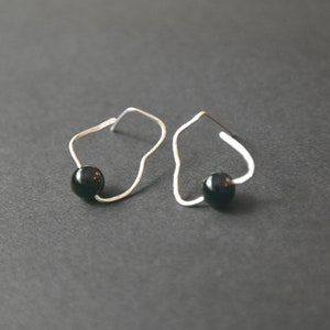 May include: A pair of silver-toned earrings with a unique, abstract design. Each earring features a small, glossy black bead suspended within a curved, open frame. The earrings are displayed against a dark gray background.