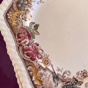 May include: A close-up of a decorative, off-white plate with a scalloped edge. The plate features a brown border with floral and botanical designs in shades of pink, blue, yellow, and green. The plate has a vintage aesthetic.