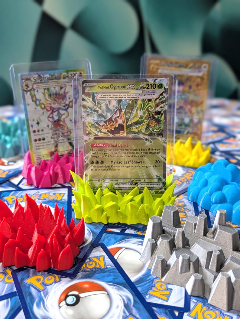 May include: Four 3D printed card holders in different colors and designs. The holders are made to display trading cards. The holders are red, yellow, gray, and blue. The holders are on a table with a variety of trading cards.