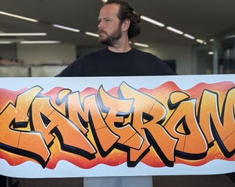 May include: A large rectangular white poster featuring the name CAMERON written in bold graffiti-style lettering. The letters are colored in shades of orange and yellow with black outlines, set against a vibrant red and orange spray-painted background.