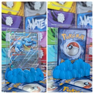 May include: A blue plastic stand with a rounded, cloud-like design holds a clear plastic card holder with a Pokemon card inside. The card features a blue Blastoise Pokemon with the text "Blastoise EX" and "330".