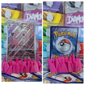May include: Two Pokemon trading cards displayed on pink plastic card holders. The card on the left is a Lileep card with a light pink background and a picture of a Pokemon with a flower on its head. The card on the right is a Pokemon card with a blue background and a picture of a Pokeball.