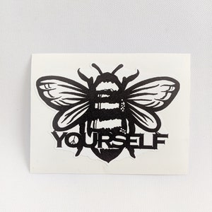 May include: A black and white sticker featuring a stylized bee design. The bee has detailed wings and body markings. The word "YOURSELF" is printed in a bold font below the bee, with a peace sign replacing the "O".