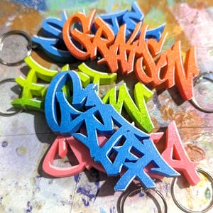 May include: Five colourful keychains with different names written in a graffiti style. The keychains are made of foam and have a 3D effect. The names are "Grayson", "Eliza", "Griffin", "Maya", and "Luca".