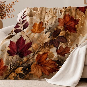 Peut inclure: Un plaid en peluche arborant un imprimé détaillé de feuilles d'érable d'automne dans des tons rouge profond, orange brûlé et brun doré sur un fond beige marbré. L'envers de la couverture est en tissu blanc uni à la texture douce.