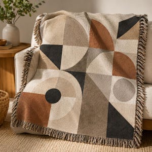 May include: A geometric patterned throw blanket in shades of beige, brown, and black, draped over a white sofa. The design features circles, triangles, and squares. The blanket has a fringed edge.