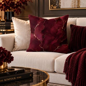 May include: A square throw pillow featuring a deep burgundy and wine-colored abstract marble pattern with thin, shimmering gold veins running throughout the design. The pillow is displayed on a light-colored sofa alongside other cushions.