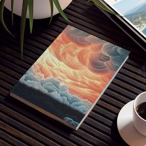 May include: A notebook with an abstract cloud design in orange, yellow, and blue tones sits on a dark brown slatted table. A white coffee cup and saucer, a potted plant, and a travel magazine are also on the table.