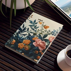 May include: A floral-covered notebook and a travel magazine rest on a dark wooden table. A white coffee cup and saucer sit nearby. A potted plant is in the background. The magazine has the word "TRAVEL" on the cover.