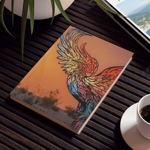 May include: A journal with a colorful phoenix design, a travel magazine, and a cup of coffee on a dark wooden table. The journal features a sunset background with a bird illustration. A potted plant is also visible.