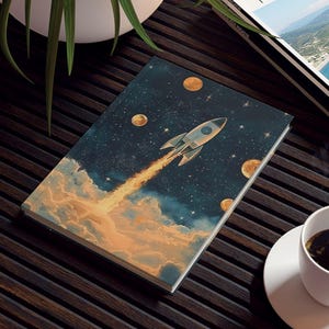 May include: A small, rectangular art print featuring a rocket ship blasting off into space, with orange clouds and golden planets. The print is on a dark wooden table, next to a cup of coffee and a travel magazine.