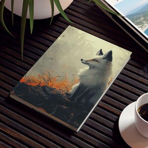 May include: A journal with a fox illustration, a travel magazine, and a cup of coffee on a dark brown slatted table. The journal features a white fox with a gray and black face. The travel magazine has the word "TRAVEL" on the cover.