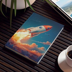 May include: A notebook with a rocket ship illustration, a travel magazine, and a cup of coffee on a dark brown wooden table. The rocket ship is red, white, and gray, with orange flames. The magazine has a landscape photo on the cover.