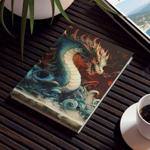 May include: A journal with a dragon illustration in shades of blue, red, and white, resting on a dark wooden table. A white coffee cup and a travel magazine are also on the table. A potted plant is in the background.