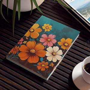 May include: A floral-themed journal with orange, pink, and white flowers on a teal background. A cup of coffee sits on a white saucer next to the journal. A travel magazine is also visible.