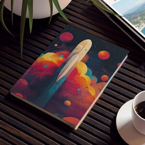 May include: A colorful notebook with an abstract design featuring a white shape and vibrant red, orange, and yellow orbs. A cup of coffee sits on a white saucer next to the notebook. A travel magazine is also on the table.