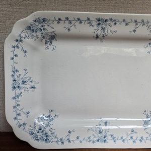 May include: A rectangular white ceramic serving platter with rounded corners, featuring a delicate blue floral vine pattern that borders the entire edge of the dish. The center of the platter is plain white, and the edges have a scalloped design.