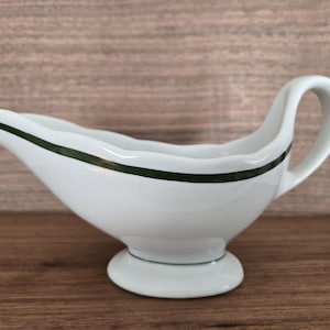 May include: White ceramic gravy boat with a dark green stripe around the rim. The gravy boat has a handle and a spout for easy pouring. The boat sits on a small, round base. The background is a wooden surface.