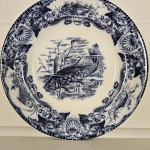 May include: A round ceramic plate featuring a blue and white transferware pattern. The center depicts a fish swimming among reeds. The wide rim is decorated with a repeating motif of smaller fish, aquatic plants, and shell-like floral elements.