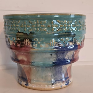 May include: A ceramic planter with a teal glaze and a decorative pattern around the top. The planter has a wide base and a narrow opening. The glaze is faded in some areas, revealing a multi-colored pattern underneath.