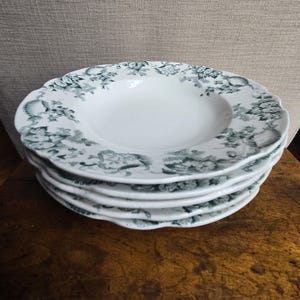May include: A stack of five white ceramic soup bowls with a scalloped edge. Each bowl features a floral and seashell pattern in shades of green around the rim. The bowls are stacked on a wooden surface.