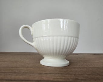 May include: Cream-colored teacup with a handle and a pedestal base. The cup has a ribbed design on the lower portion and a smooth upper section. The teacup is made of ceramic and is sitting on a wooden surface.