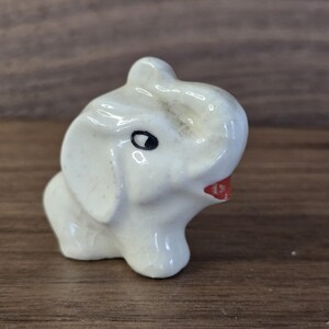May include: A small, white ceramic elephant figurine with a red mouth and black eyes. The elephant has large ears and a trunk that is slightly raised. The figurine is on a wooden surface.
