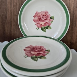 May include: A stack of white ceramic plates with a green border and a pink rose design in the center. The plates are stacked on top of each other, with two plates visible in the image. The plates are on a white surface.