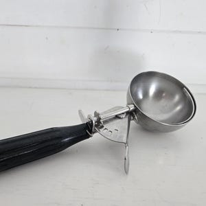 May include: Vintage ice cream scoop with a stainless steel bowl and mechanical release. The handle is black with a white tip. The scoop is designed for portioning ice cream and other frozen desserts.