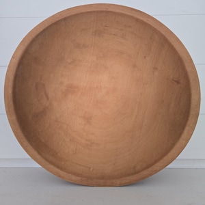 May include: A large, round, wooden bowl with a smooth, matte finish and a natural light brown color. The bowl features a wide, open interior with visible wood grain patterns and a thick, rounded rim, showcasing a simple, functional design.