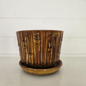 May include: A cylindrical ceramic planter with a glossy, metallic bronze glaze and a vertical bamboo-like relief pattern. The planter sits on a matching circular saucer with a slightly lighter, mottled brown finish and a raised rim.