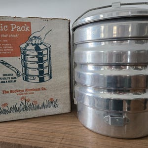 May include: Vintage aluminum picnic pack with a matching box. The box reads "Picnic Pack" and includes text about its uses. The stackable container includes 4 utility pans and a skillet. The Buckeye Aluminum Co. product is designed for family picnics and beach parties.