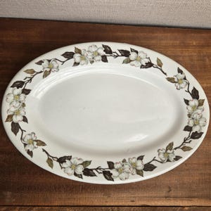 May include: An oval-shaped white ceramic serving platter featuring a continuous border pattern of white dogwood flowers with yellow centers and dark brown leaves arranged along the rim of the dish, creating a decorative floral frame.