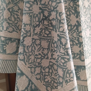 Puede incluir: Un mantel floral turquesa y blanco drapeado sobre una mesa redonda. La tela presenta un patrón repetido de flores y follaje estilizados. El borde tiene un diseño a rayas.