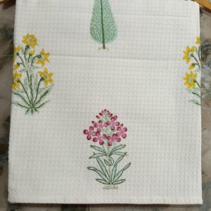 May include: A white waffle-weave fabric featuring a repeating block-print pattern. The design includes a central pink floral motif, a green tree above it, and yellow floral clusters on the left and right sides, all with green stems and leaves.