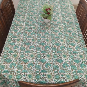 Pode incluir: Uma toalha de mesa branca com um padrão floral verde e marrom. A toalha de mesa está posta sobre uma mesa de madeira com quatro cadeiras. Um pequeno buquê de flores cor-de-rosa está no centro da mesa.