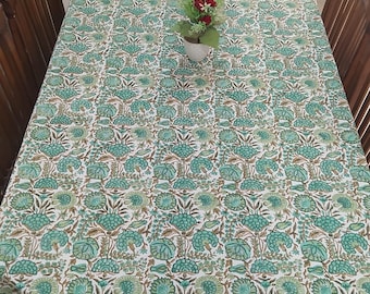May include: A white tablecloth with a green and brown floral pattern. The tablecloth is laid on a wooden table with four chairs. A small bouquet of pink flowers is in the center of the table.