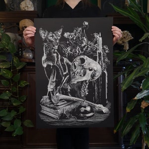May include: A black and white print of a still life featuring a black cat, a skull, a candle, and flowers. The print is on black paper with silver ink.