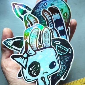 May include: A set of holographic stickers featuring various cartoon characters, including a skull with bunny ears, a bird, and a cat. The stickers are iridescent and have a blue and green color scheme.