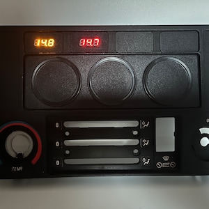 Könnte beinhalten: Schwarzes Kunststoff-Armaturenbrett mit einem digitalen Display, das "148" und "083" anzeigt, drei runden Knöpfen, einem Temperaturregler mit der Beschriftung "TEMP" und einem Lüftersteuerungsfeld mit drei Schiebereglern und einem Knopf.