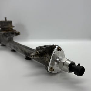 Può includere: Un gruppo di tubi carburante grigio metallizzato con una piastra argentata e un connettore nero. Il gruppo ha vari raccordi e bulloni, su uno sfondo bianco.