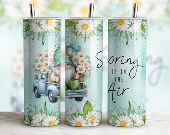 Op de afbeelding: Een turquoise blauwe tumbler met een witte dwerg die een mand met witte bloemen draagt in een blauwe pick-up truck. De tumbler heeft een wit madeliefje ontwerp en de tekst "Spring is in the air".