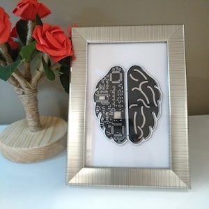 Pode incluir: Uma obra de arte emoldurada com um design de cérebro estilizado. Metade do cérebro é um padrão de placa de circuito, a outra é cinza escuro liso. A moldura prateada envolve a obra de arte, que está sobre um fundo branco.