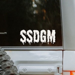 Peut inclure: Un autocollant en vinyle blanc avec le texte "SSDG" en lettres qui coulent, appliqué sur la lunette arrière d'un SUV gris.