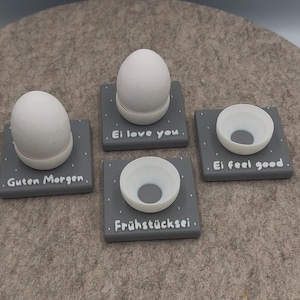 Puede incluir: Cuatro hueveras grises con huevos blancos o hueveras. Cada soporte tiene lunares blancos y una frase: "Guten Morgen", "Ei love you", "Ei feel good" y "Frühstücksei". Los huevos y las tazas son blancos.