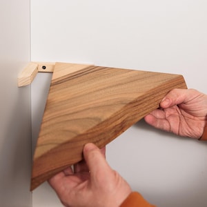 Peut inclure: Une étagère triangulaire en bois est en cours d'installation sur un mur blanc. L'étagère est en bois brun clair avec un motif de grain naturel. L'étagère est maintenue en place par les mains d'une personne.