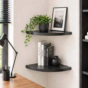 Op de afbeelding: Twee zwarte zwevende hoekplanken gemonteerd aan een witte muur. De bovenste plank bevat een groene plant in een pot en een ingelijste zwart-wit print van een berglandschap. De onderste plank bevat vier boeken met witte en zwarte ruggen en een zwarte vaas.