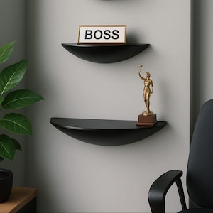 May include: Two black, semi-circular shelves on a grey wall. The top shelf has a sign reading "BOSS". The lower shelf displays a gold statue of a person holding a star. The shelves are a modern design.