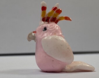 May include: A small pink and white ceramic figurine of a cockatoo with a red and yellow crest.